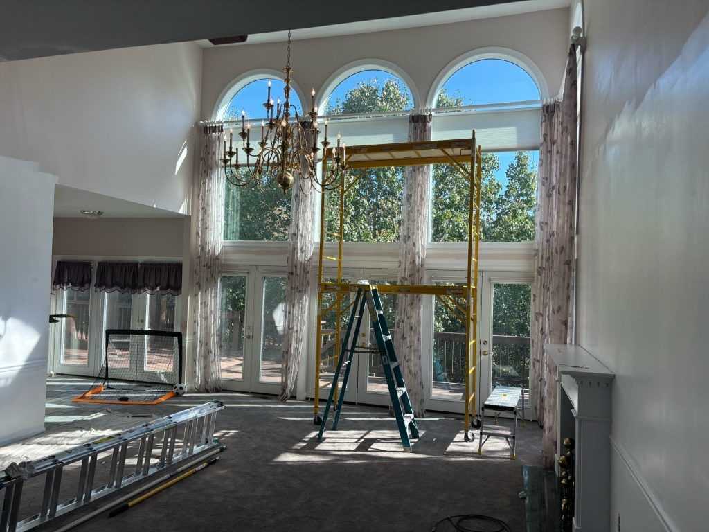 Interior view of a room with tall windows, scaffolding, and a chandelier. Sunlit space with curtains and open doors.