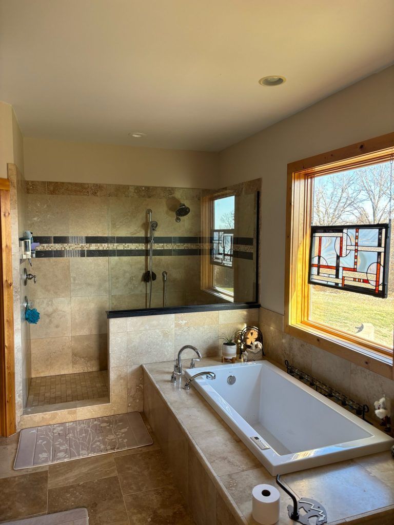 Bathroom with a walk-in shower, bathtub, and window. Light-colored stone walls. Sunny natural light.