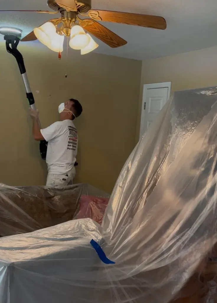 Person sanding a ceiling, wearing a mask, with plastic sheeting covering furniture in a room.