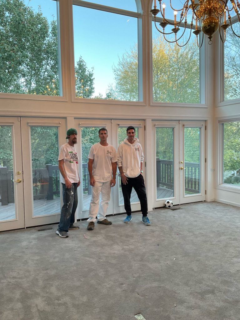 Three men stand in a room with large windows. The room has gray carpet, with trees visible outside.