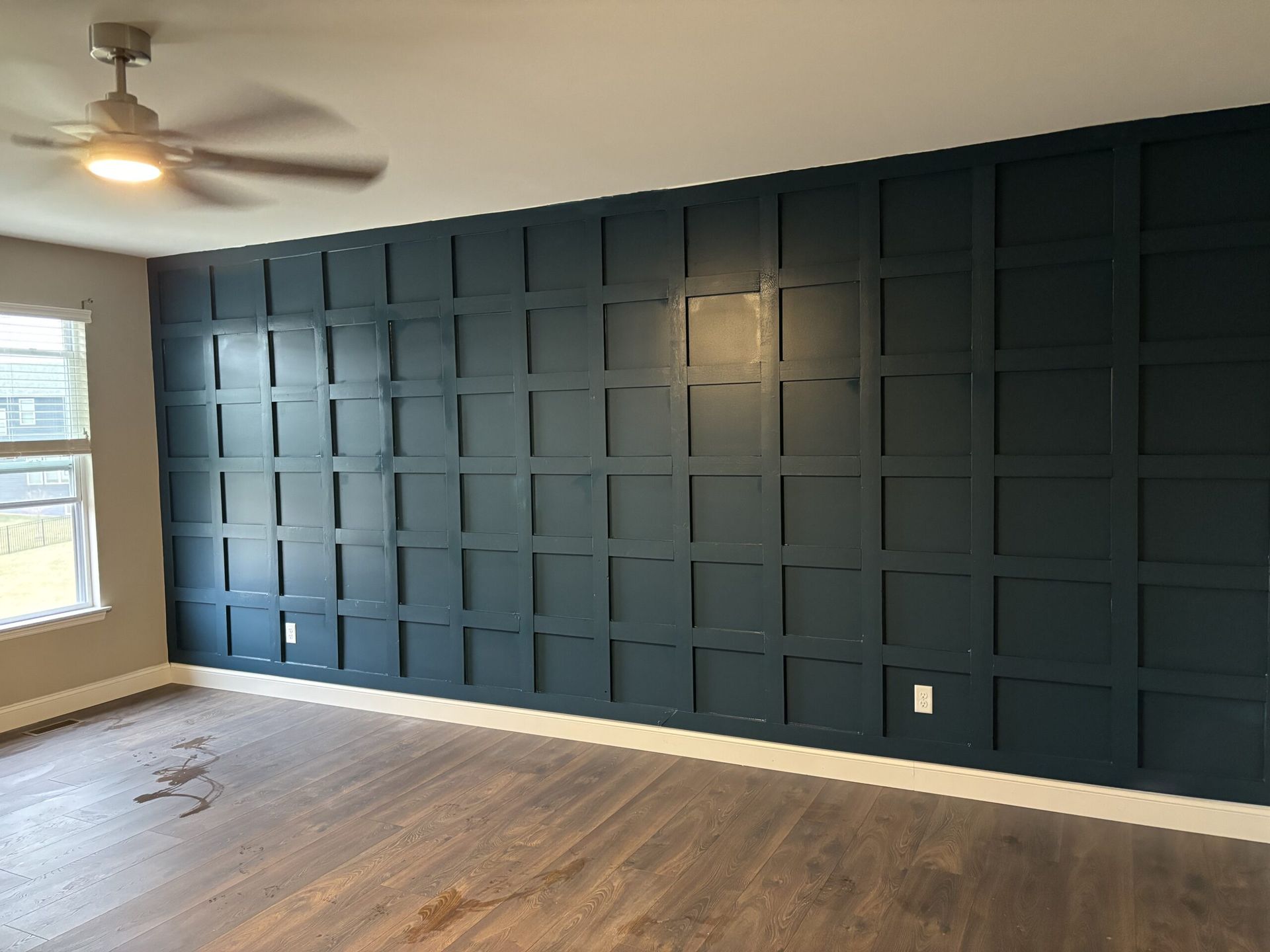Dark teal paneled accent wall in a room with wood flooring, ceiling fan, and window.