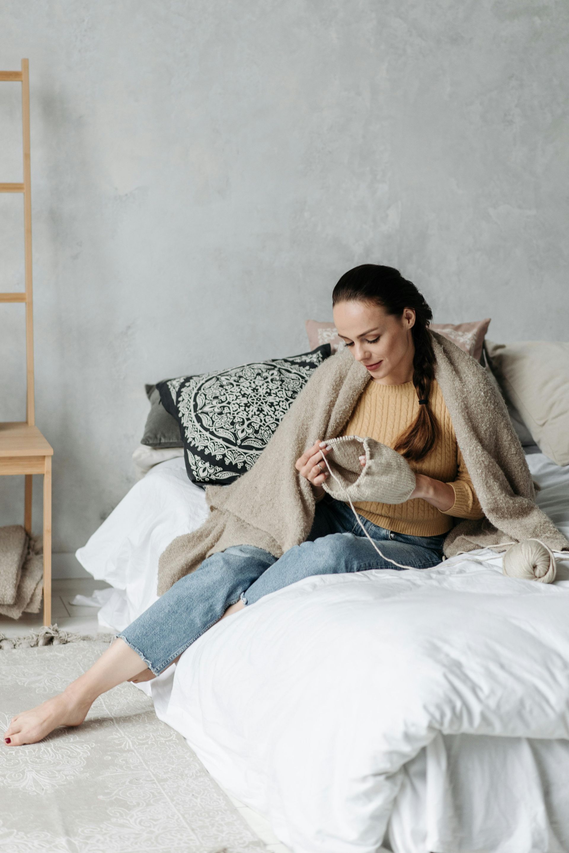 Woman knitting on a bed, wrapped in a blanket. She's smiling, sitting on white bedding with a gray wall backdrop.
