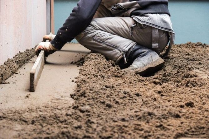 Worker leveling wet concrete beside a foundation wall with a trowel.