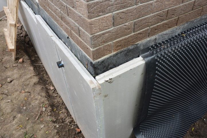 Foundation corner with white waterproofing panel and black drainage mesh against brick wall