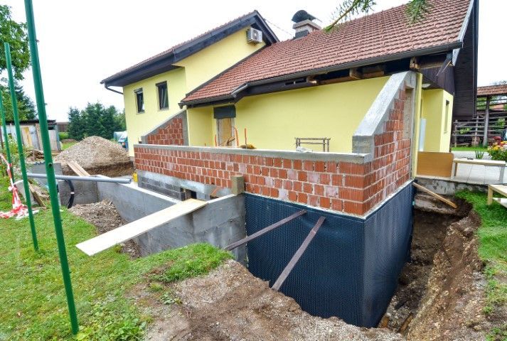 House under construction with brick foundation, yellow walls, and red roof beside an excavation trench