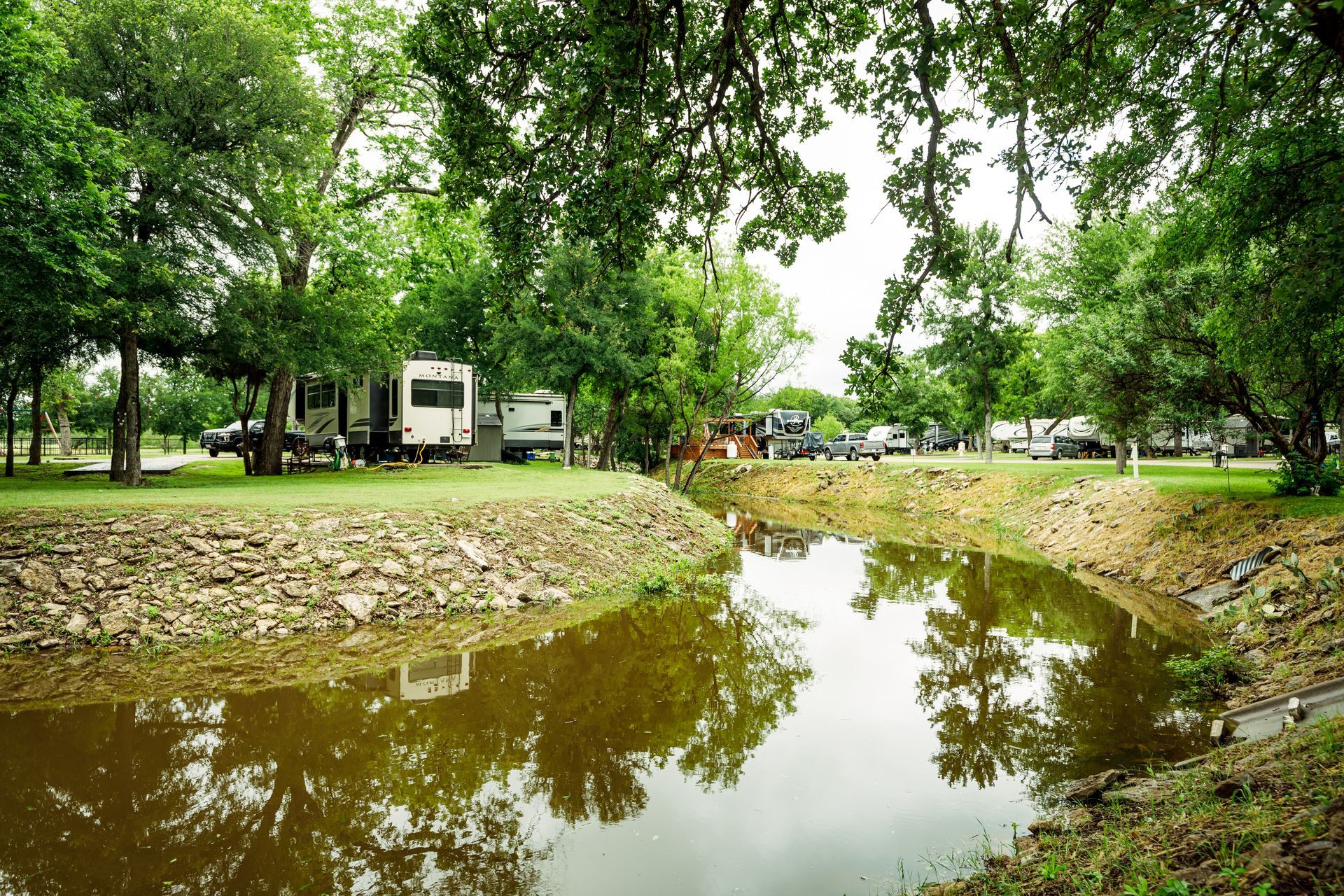A rv is parked in a campground next to a pond surrounded by trees.