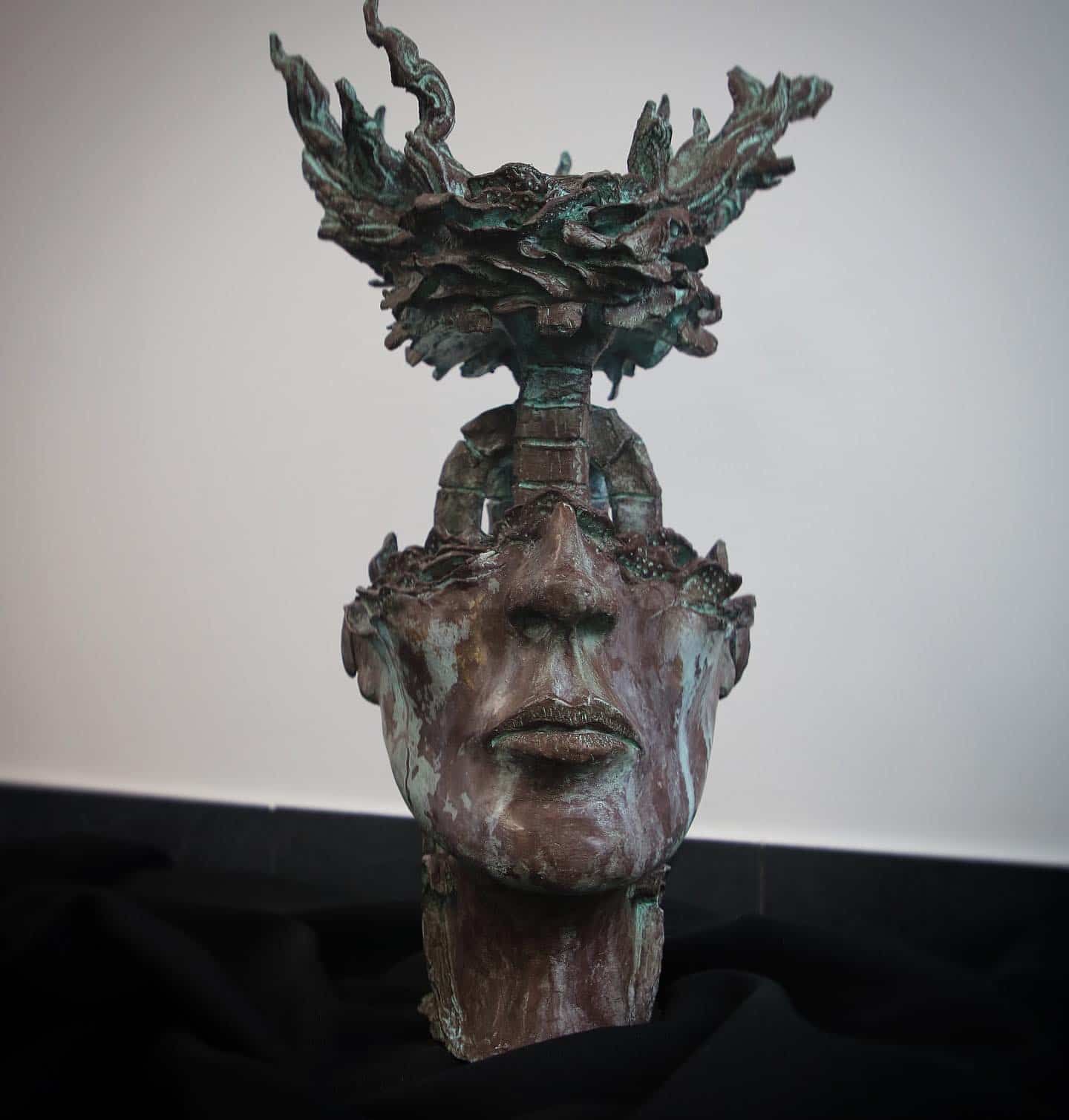 Bronze sculpture of a face topped with a stylized tree-like element; on a black cloth.