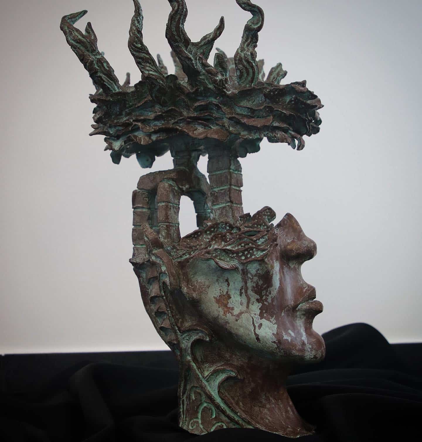 Sculpture of a face with open mouth and crown of tree-like form, dark bronze-green and brown.