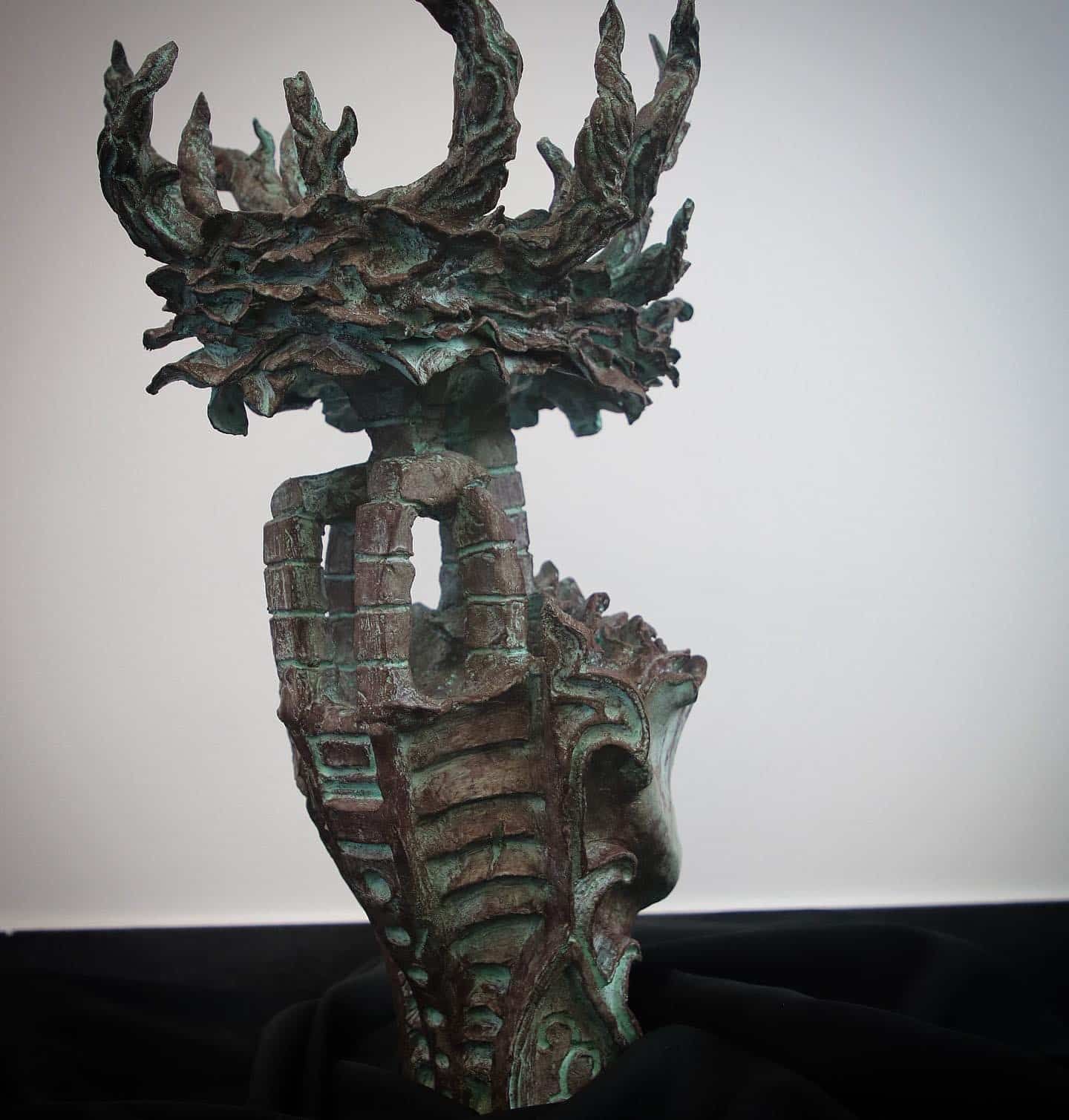 Bronze sculpture of a distressed face atop a castle structure, crowned with leafy branches.