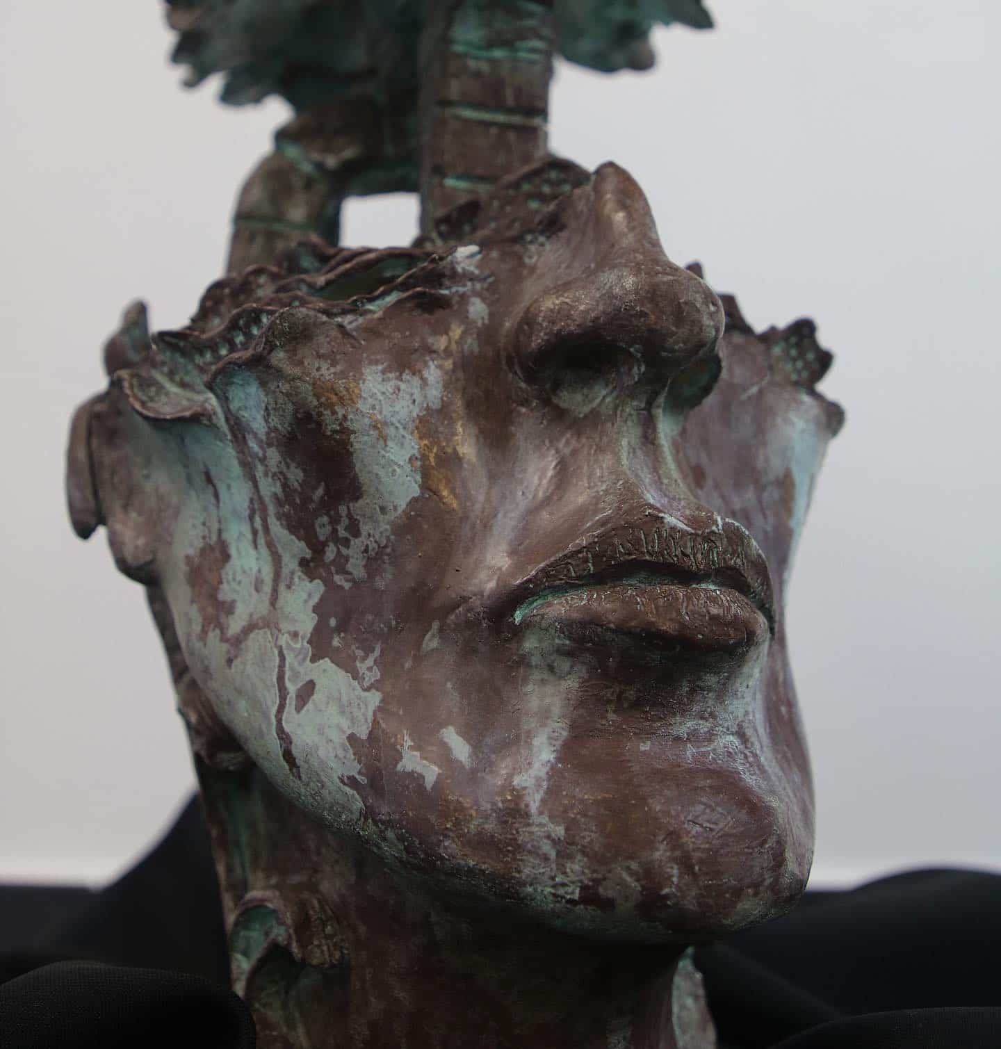 Bronze sculpture of a face with a tree growing out of the head; weathered teal and brown.