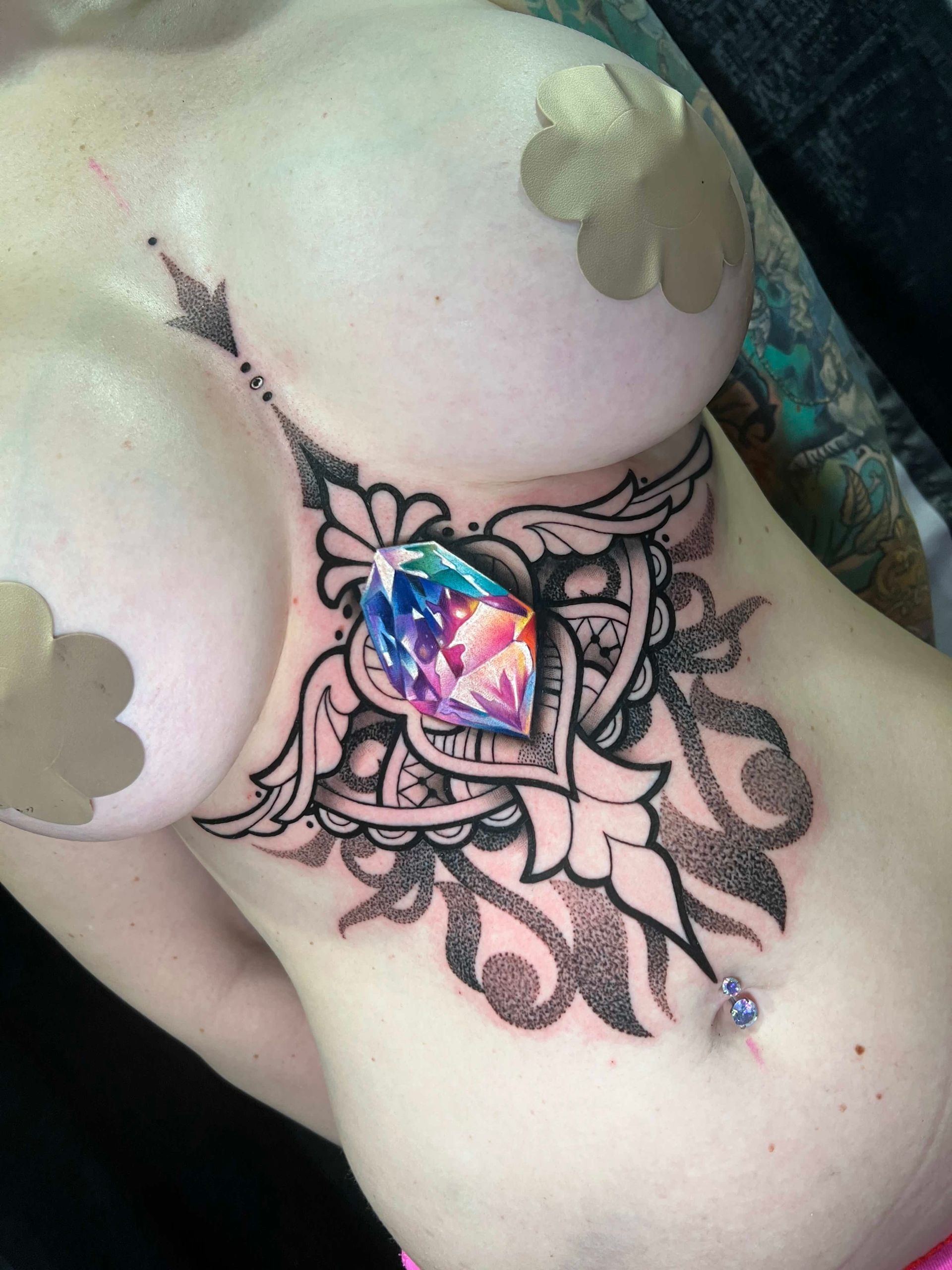 Tattoo on the chest, a large gem with floral design. Nipple pasties and navel piercing visible.