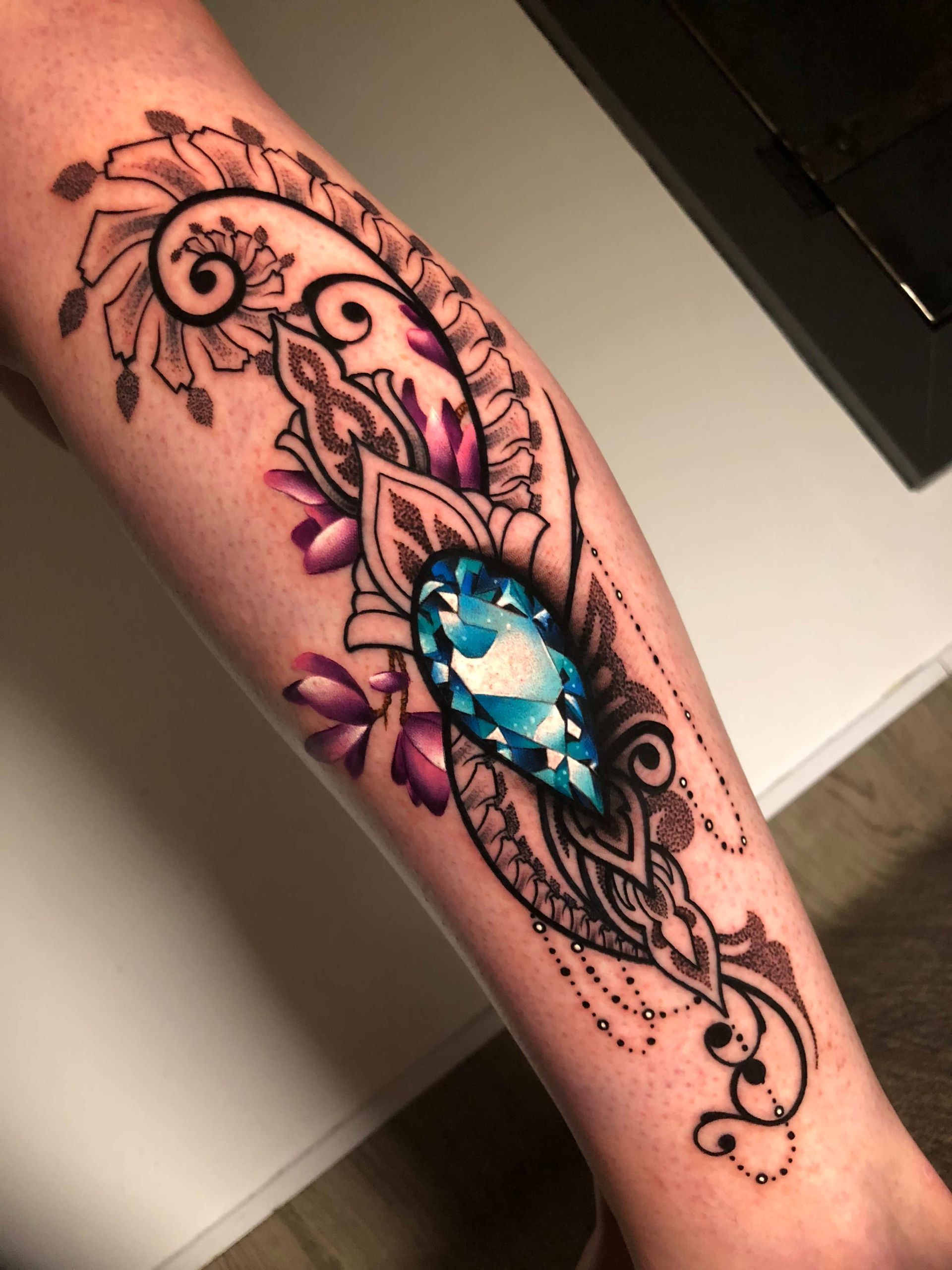 Tattoo of a large blue gemstone surrounded by black ornamental floral designs and purple flowers on a forearm.