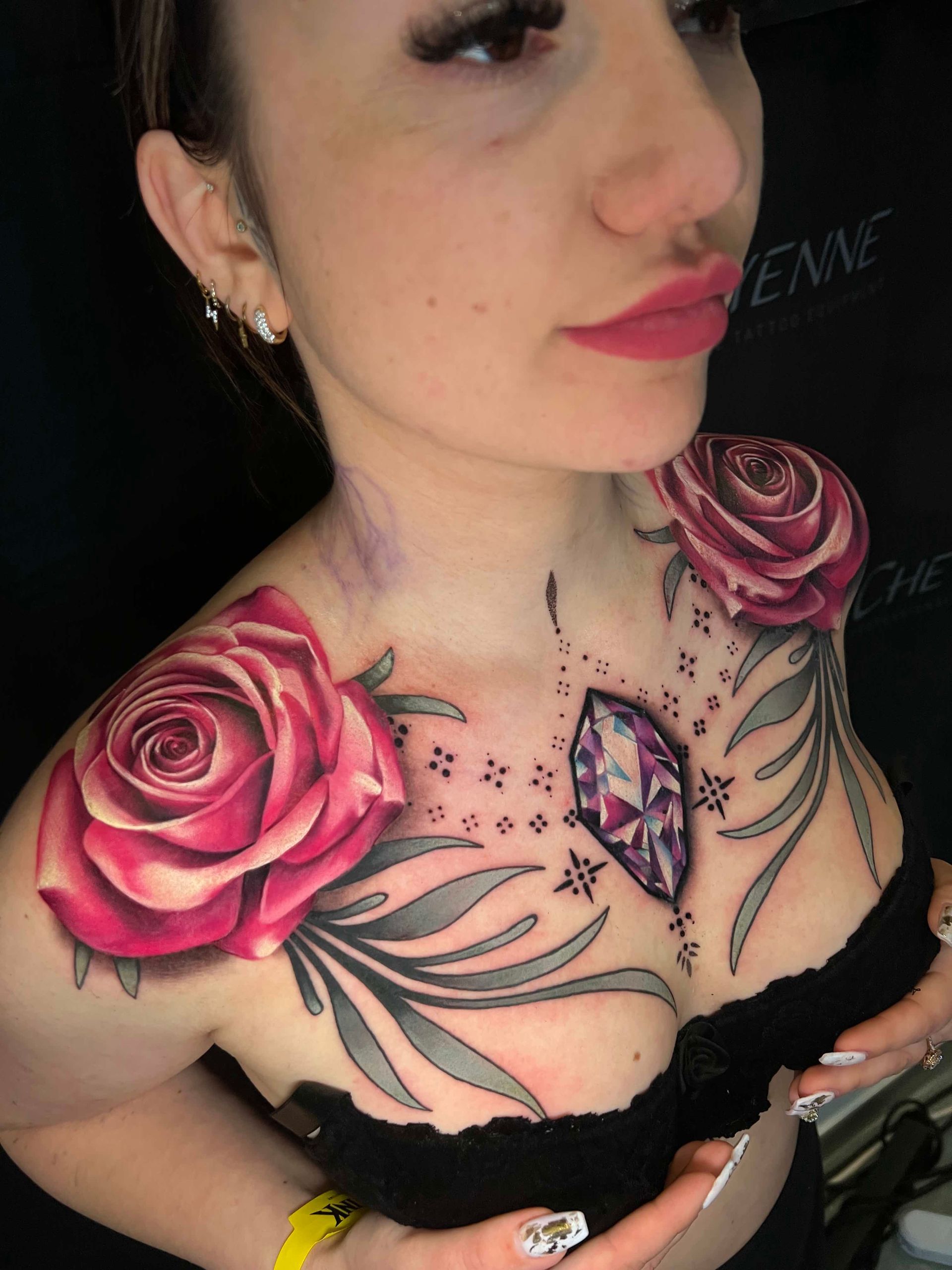 Woman with rose and gem chest tattoo. Roses are pink, gray leaves. Black background.