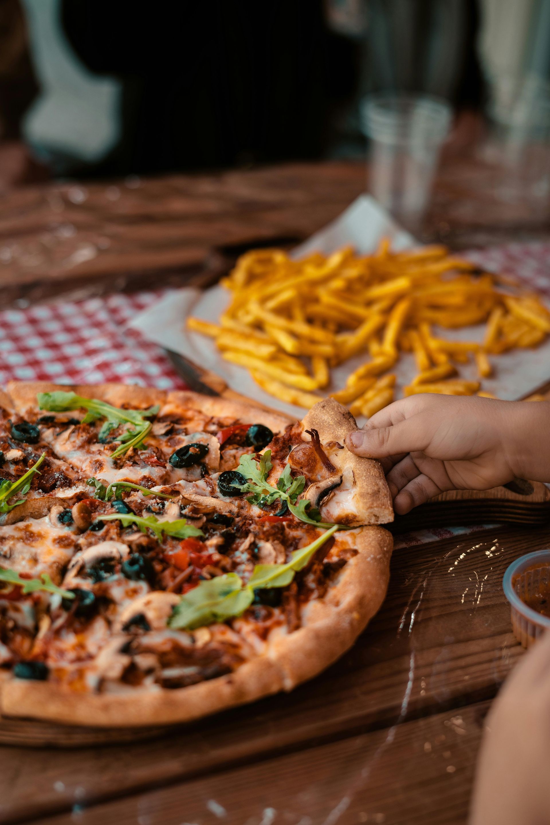 Pizza and fries on a wooden table. A hand is reaching for a pizza slice.