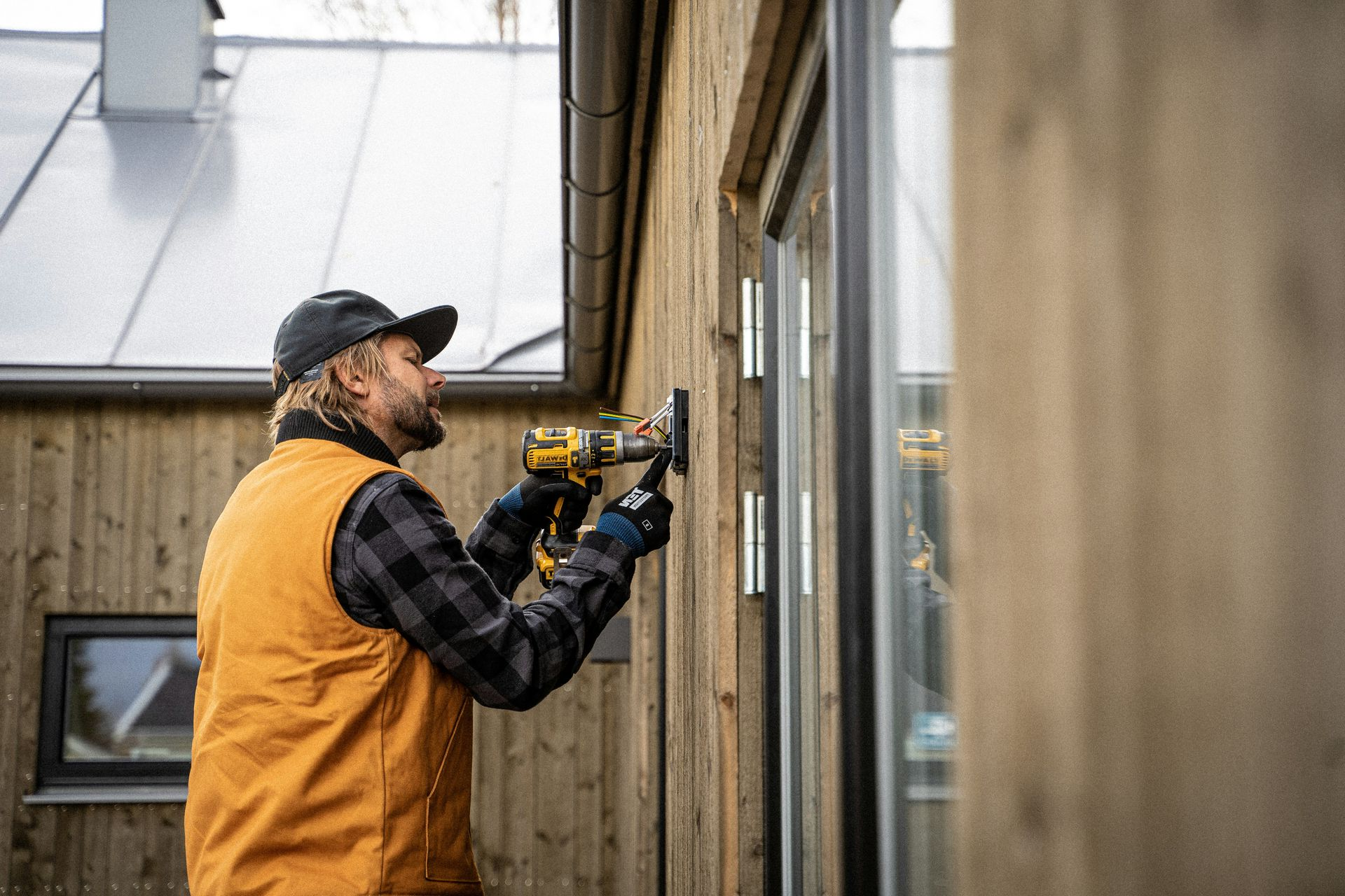 Person using a power drill on the exterior of a building with wooden siding; wearing a hat, vest, and gloves.