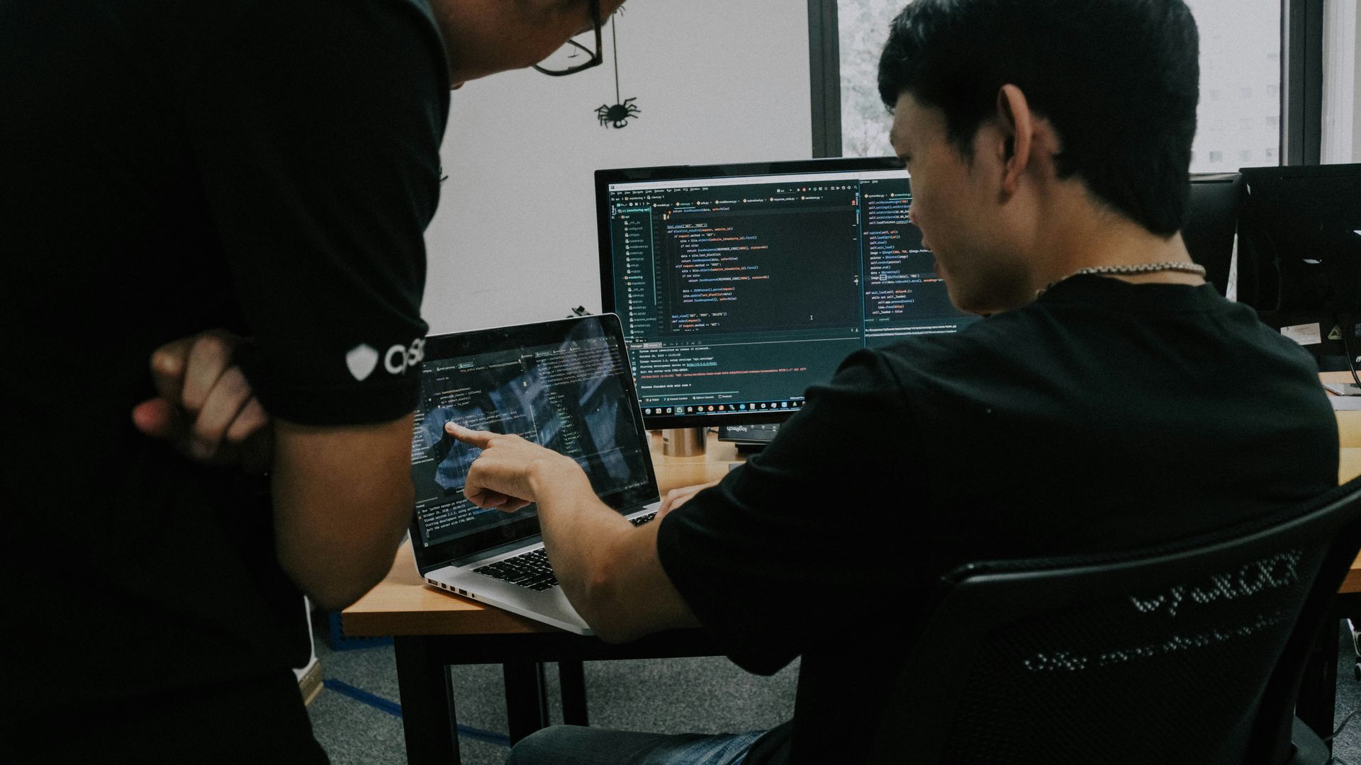 Two people collaborating at a desk, looking at a laptop screen with code, in an office.