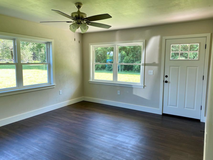Empty room with dark wood floor, three windows, white door, beige walls, and ceiling fan.