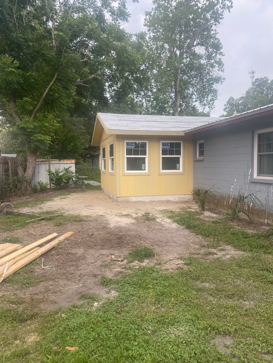 Yellow addition on a gray house with two windows. Dirt yard and trees in the background.