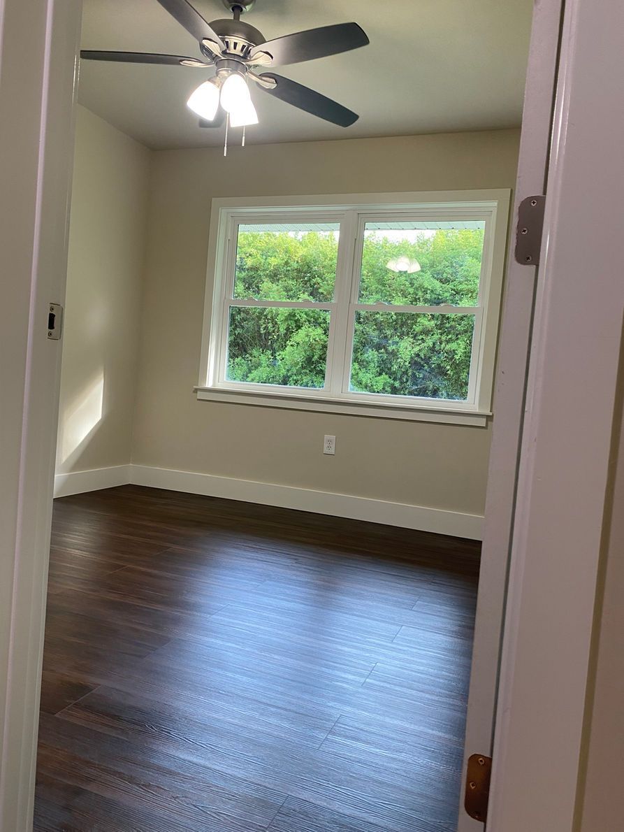 Bedroom with dark wood floor, beige walls, and white window frame.  Ceiling fan and window overlooking greenery.