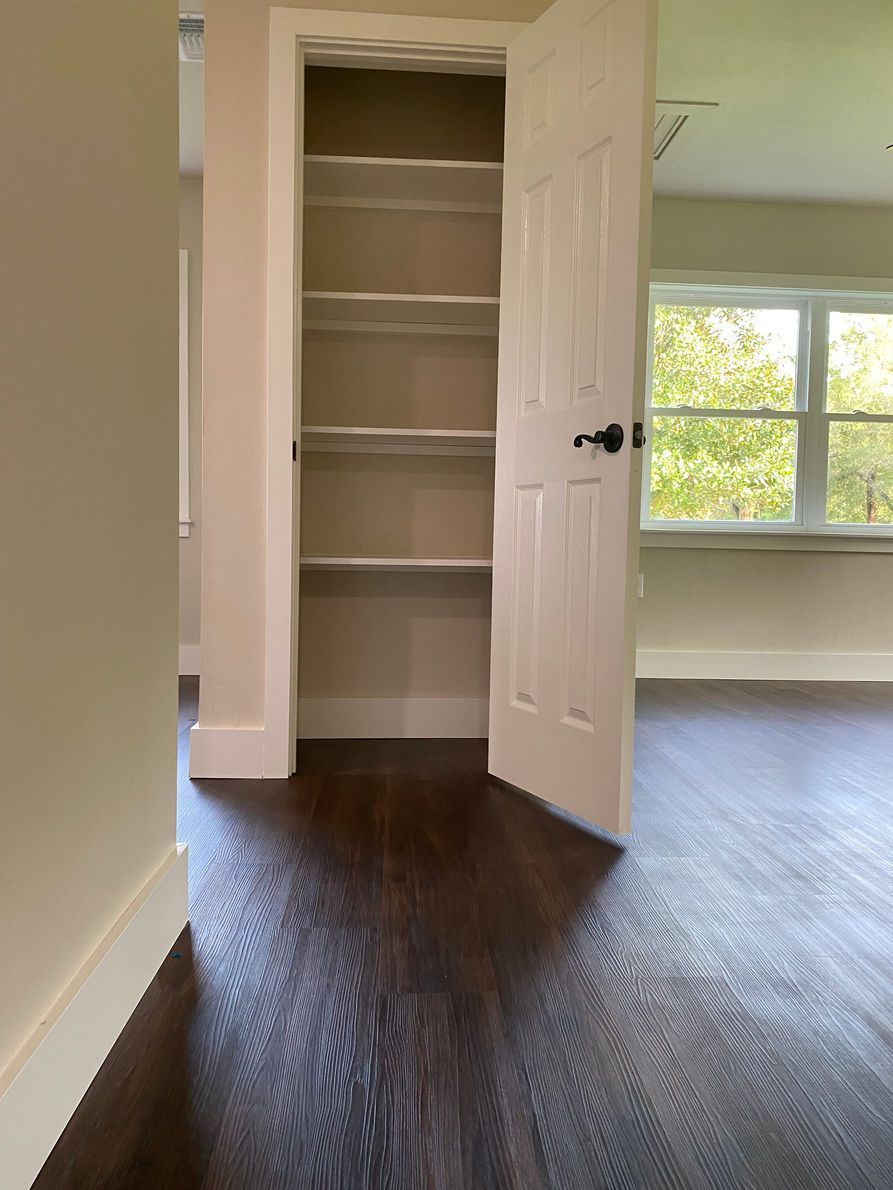 A pantry with open white door, several shelves, on dark wood floor. Window in the background.