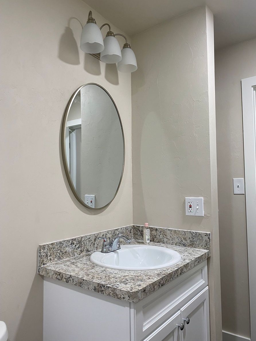 Bathroom vanity with oval mirror, sink, countertop, light fixture. White cabinet, beige walls.
