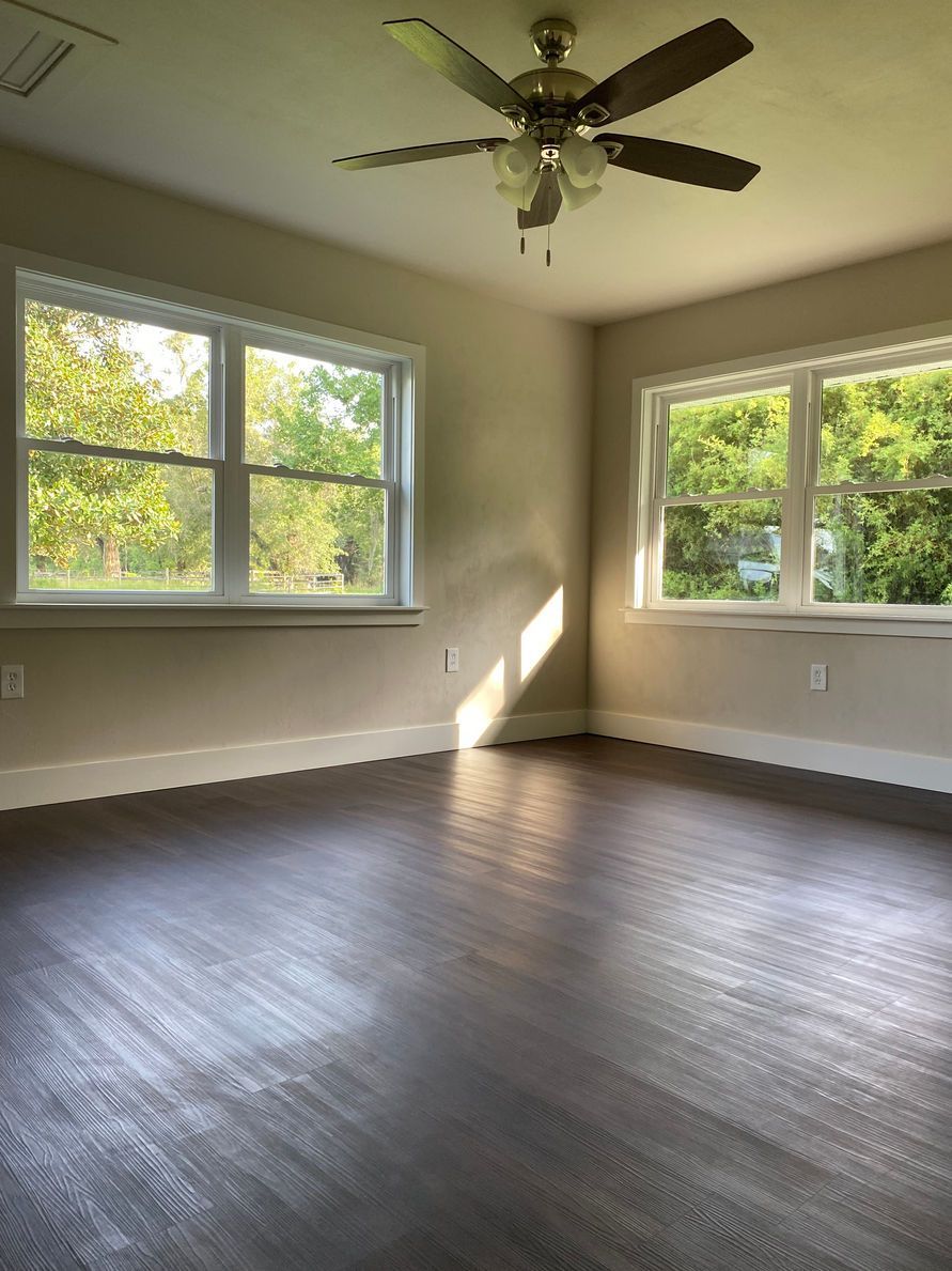 Empty room with dark wood floor, light walls, and two windows; ceiling fan.