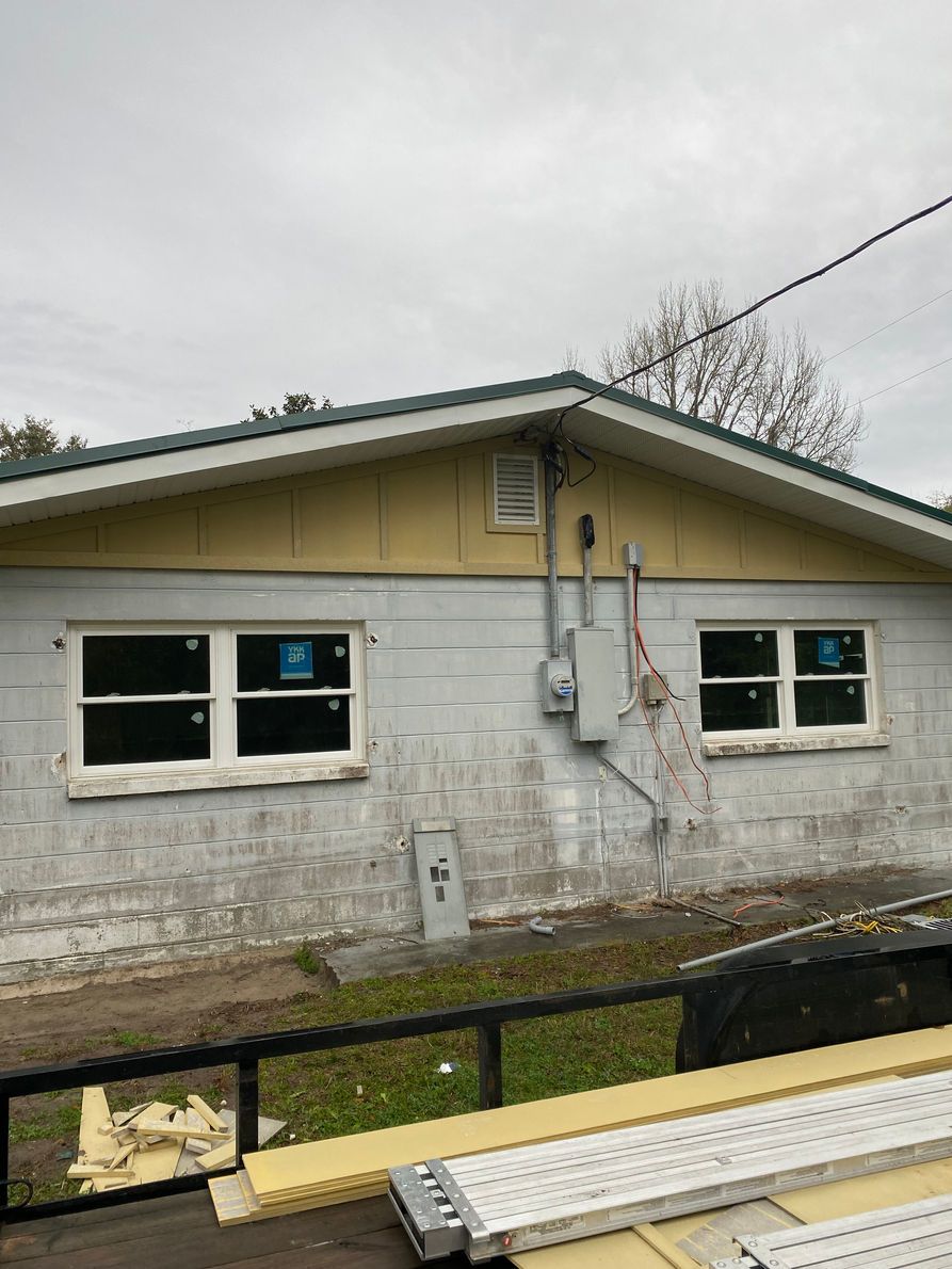 Exterior of a house with gray cinder block walls, two windows, and utility equipment. Overcast sky.