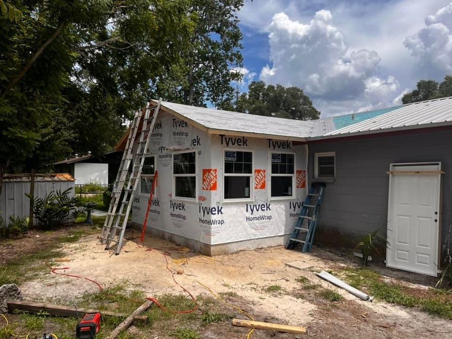 Construction of an addition to a gray building; Tyvek-wrapped walls with windows, ladders, and a sunny setting.