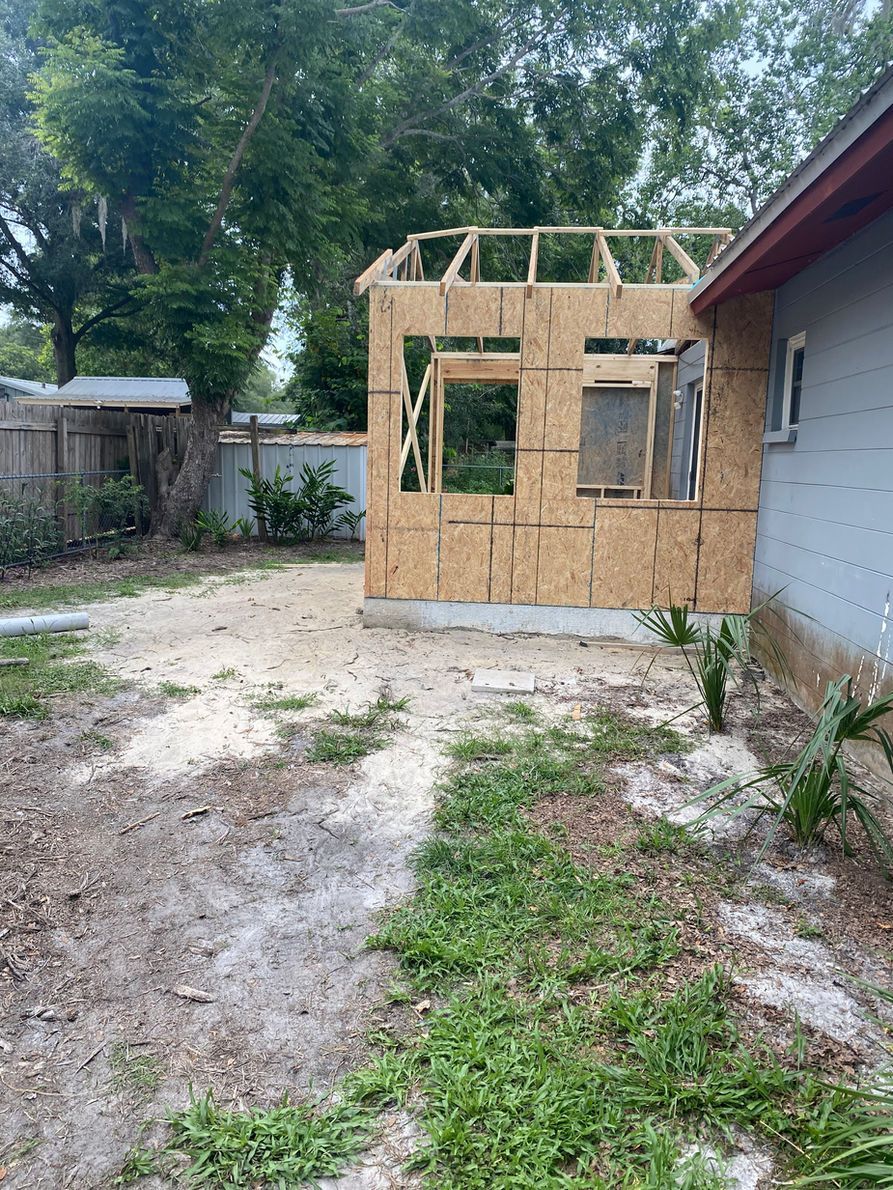 Construction of a room addition on a house; plywood walls, exposed framing, and a dirt yard.