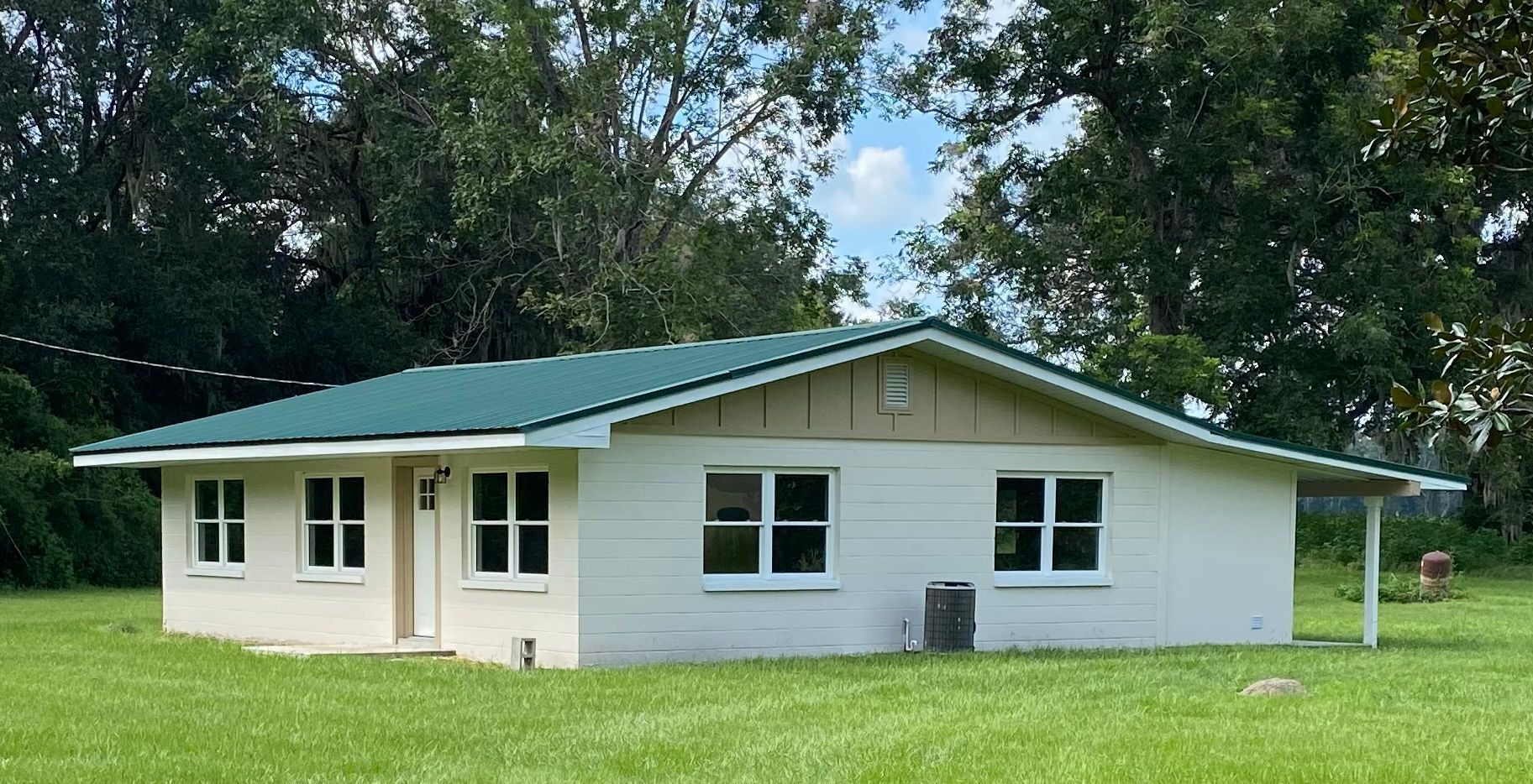 White, single-story house with green roof. Green lawn and trees in the background. Blue sky.