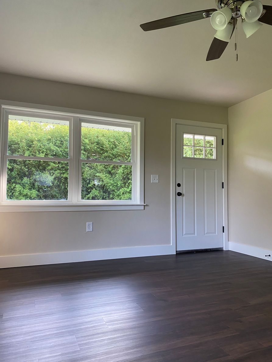 A living room with a window and door. Beige walls, white trim, dark wood floor. A ceiling fan is mounted.