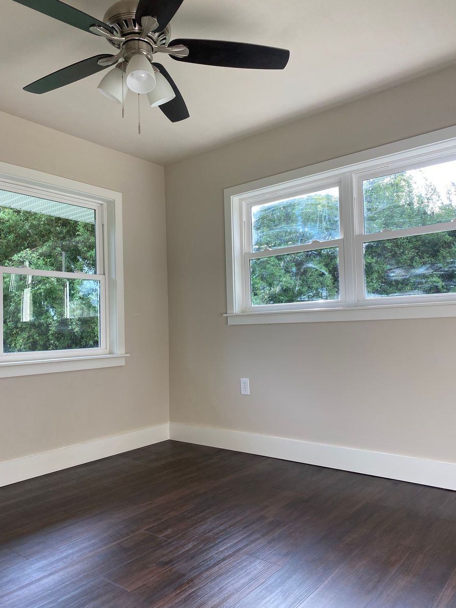 Empty room with dark hardwood floors, light beige walls, white trim, and two windows overlooking greenery.