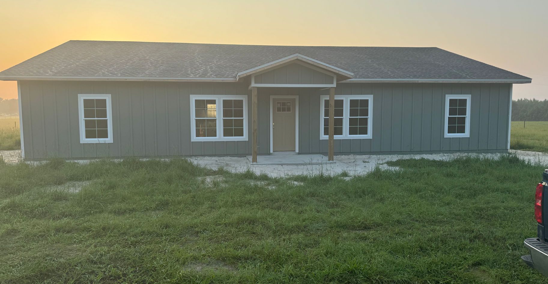 Gray house with white trim, front porch, and windows in a grassy field at sunset.