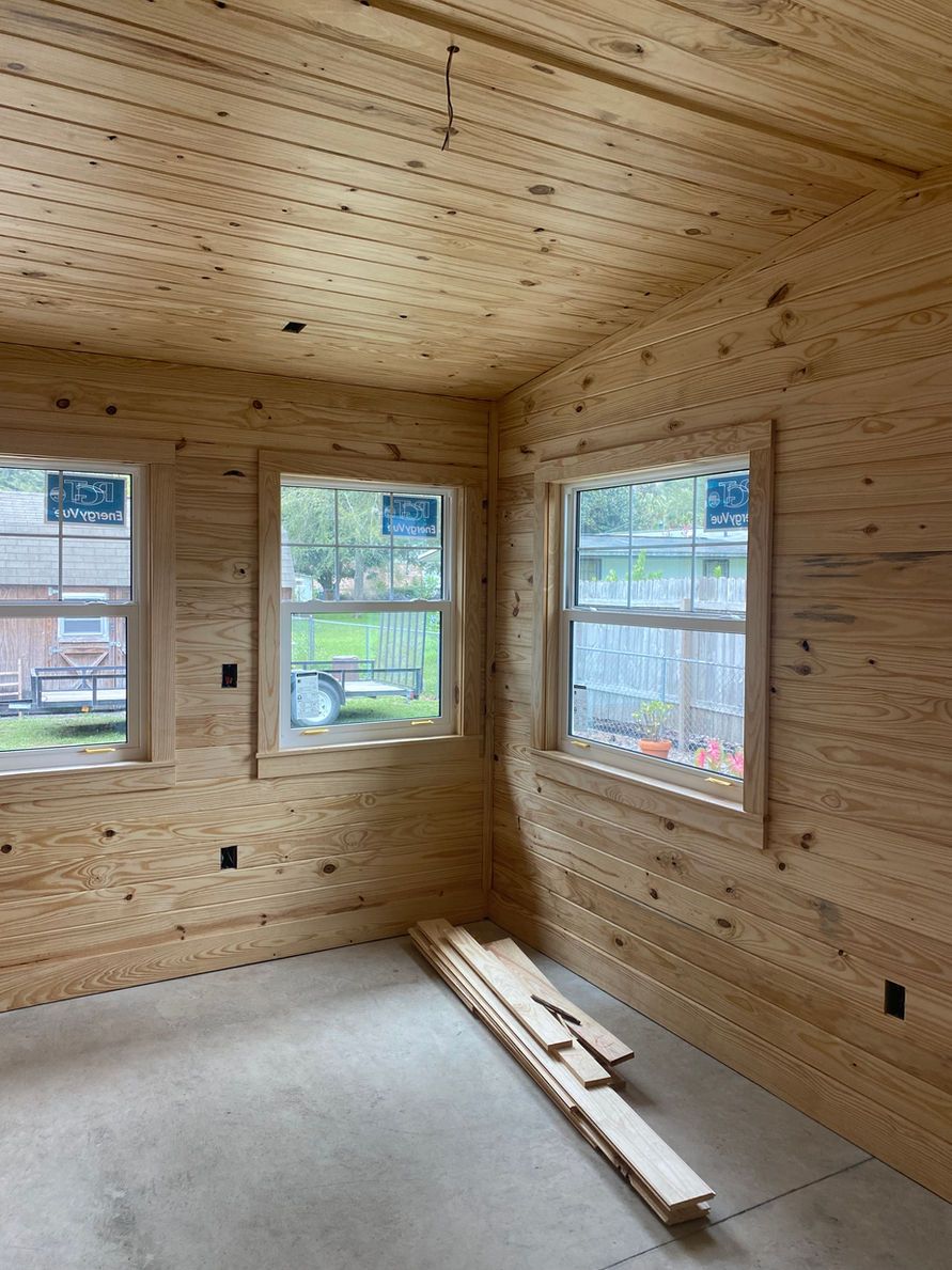 Interior of a room with wood-paneled walls and ceiling, three windows, and stacked lumber.