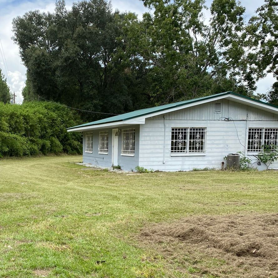 A small, light blue house with a green roof on a grassy lawn, trees in the background.