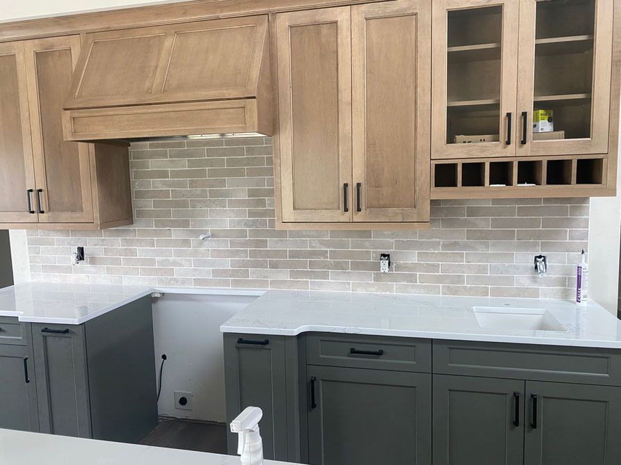 Kitchen with light wood cabinets, gray lower cabinets, white countertops, and tan brick backsplash.