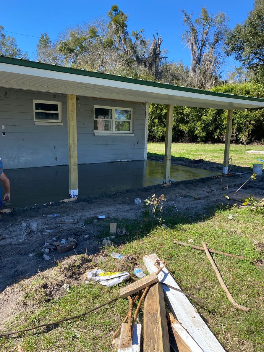 Construction site: concrete patio, gray building, green roof. Wooden support beams.