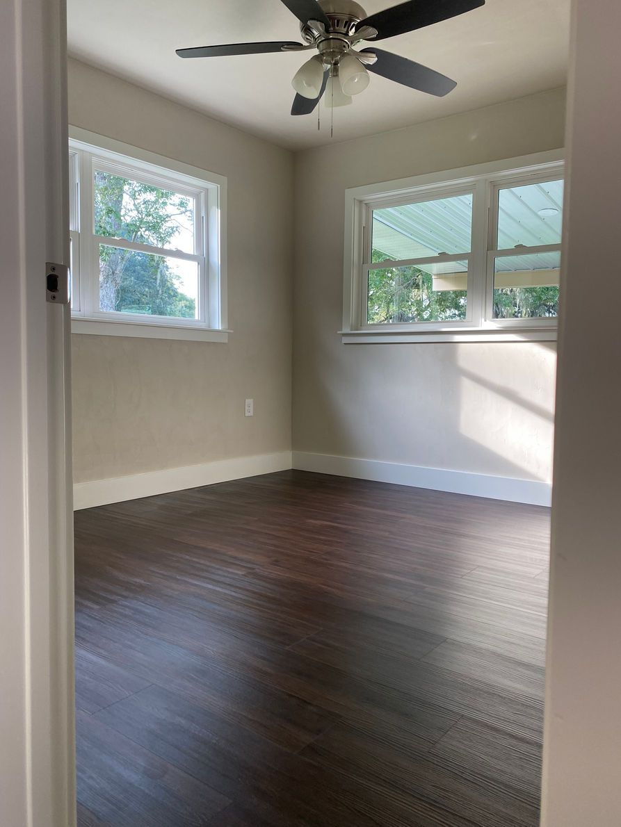 Empty bedroom with dark wood-look floor, light gray walls, two windows, white trim, and ceiling fan.