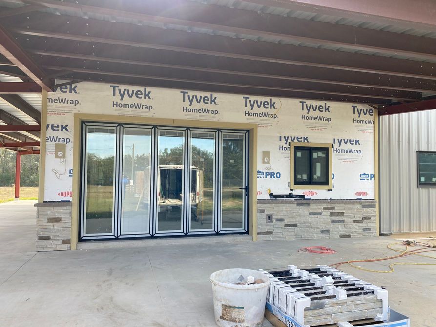 Construction site: building with Tyvek wrap, folding glass doors, stone veneer, and a small window under a metal awning.