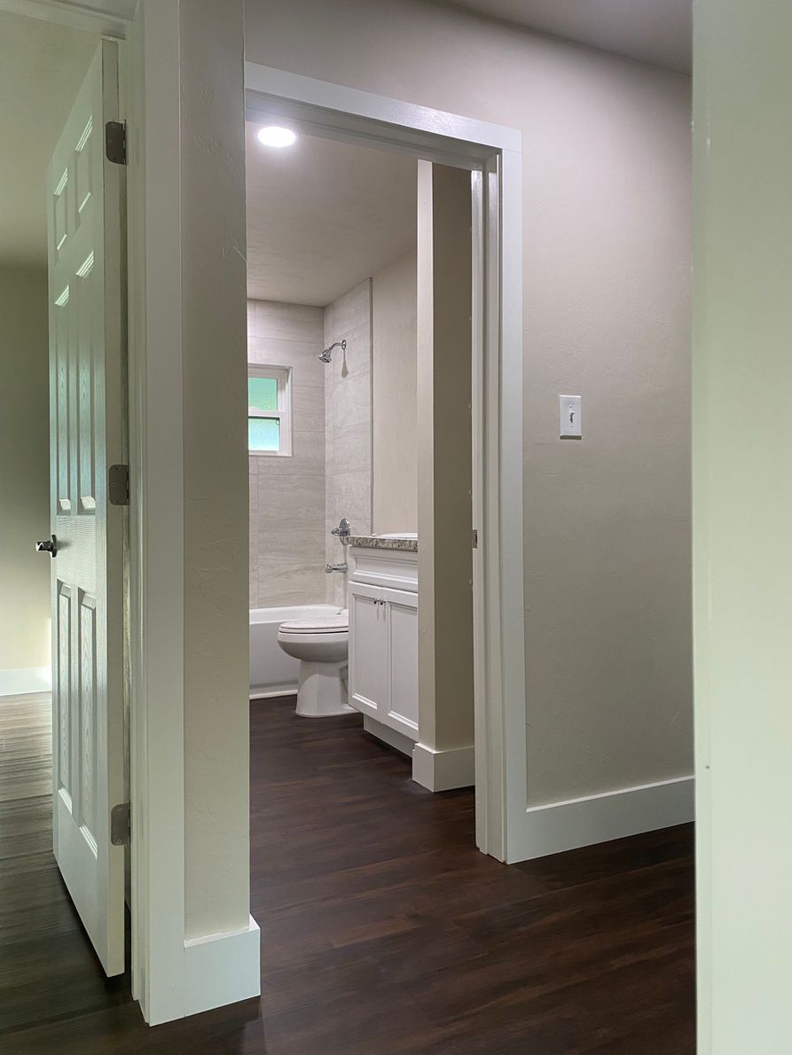 Hallway doorway leads to a bathroom with a tub, toilet, and white vanity. Dark wood flooring and white trim.