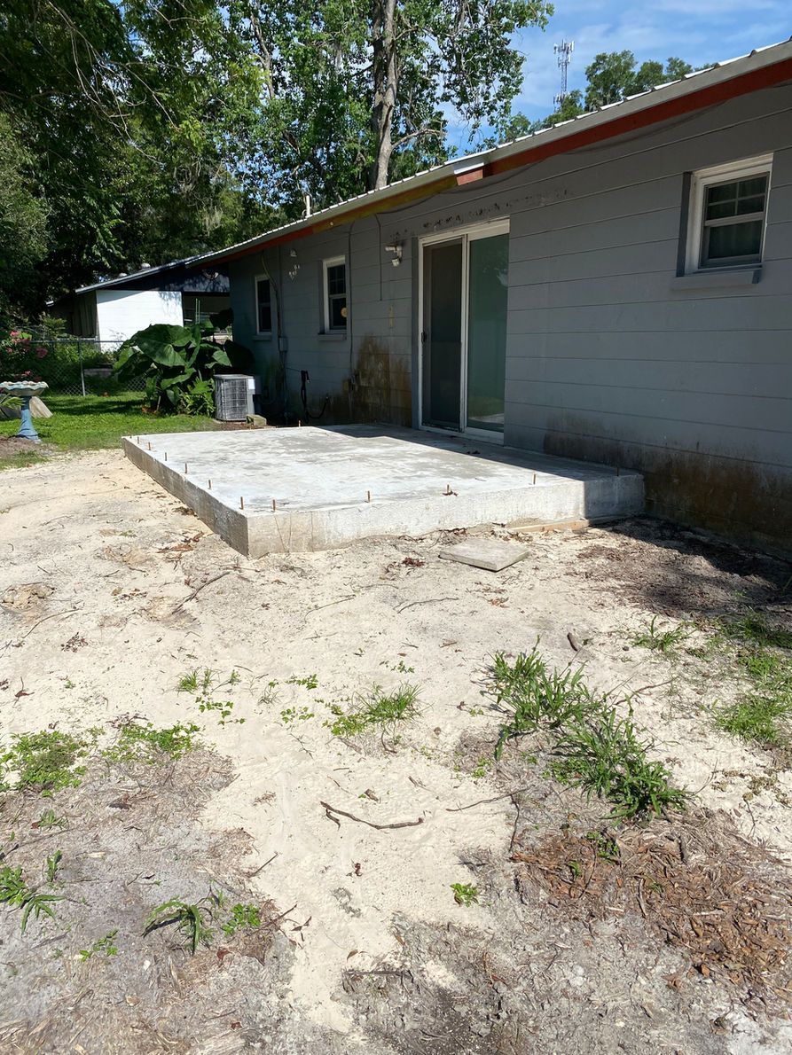 Concrete patio foundation outside a gray brick house with a sliding glass door. Dirt and sparse vegetation surround it.