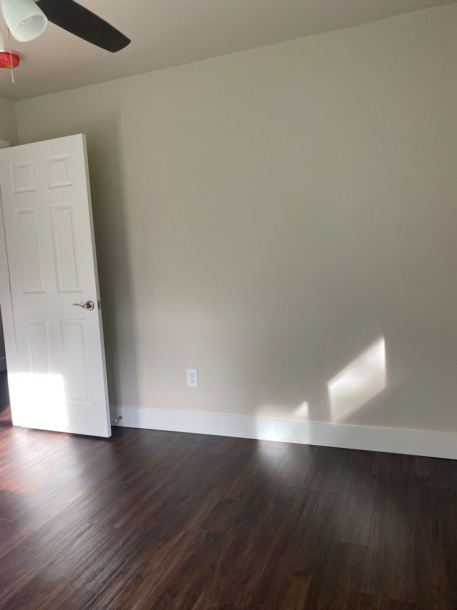 Empty room with dark wood floor, white door, and beige walls; sunlight.
