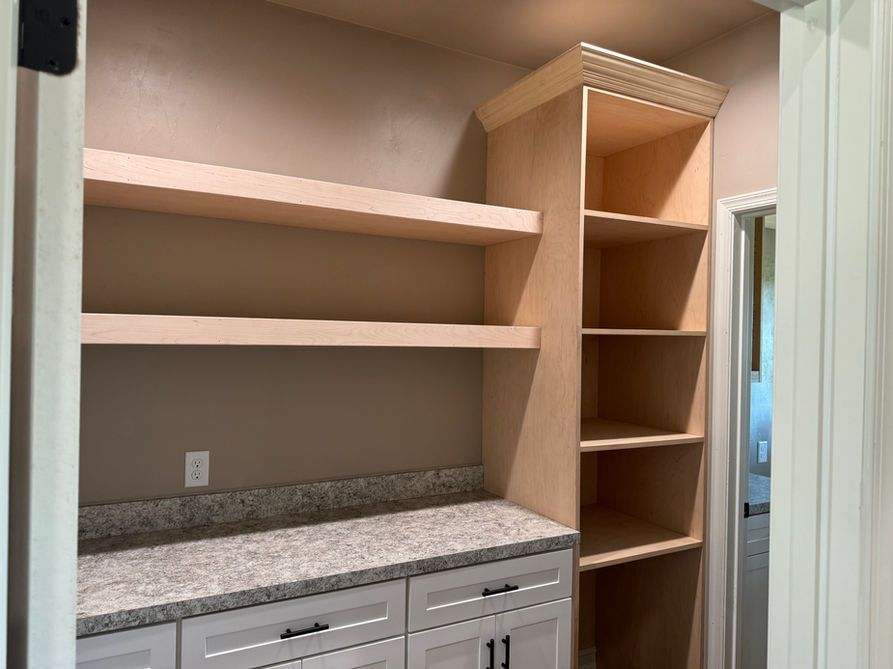 Pantry with light wood shelves, granite counter, white cabinets. Right is tall shelving; doorway is on the right.