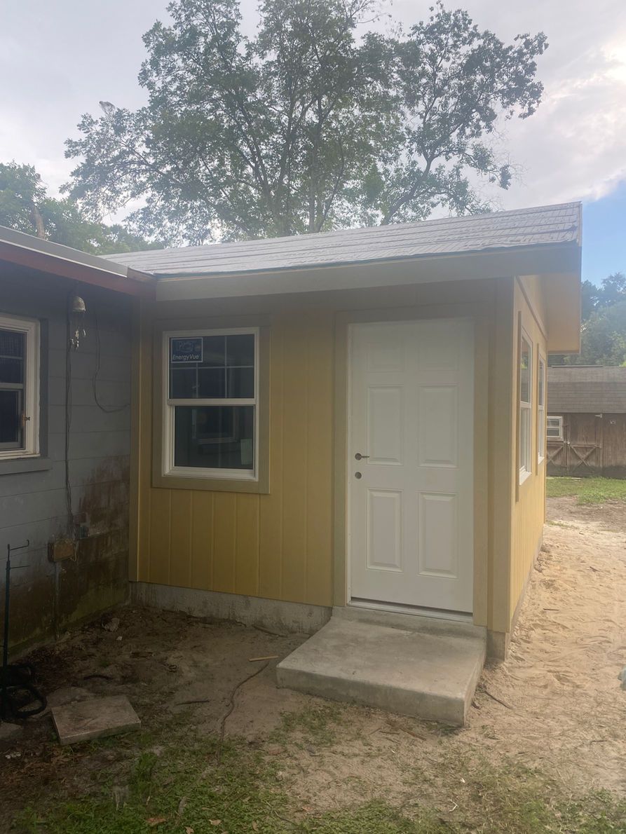 Yellow shed with white door and window, concrete step, gravel ground, next to gray building.