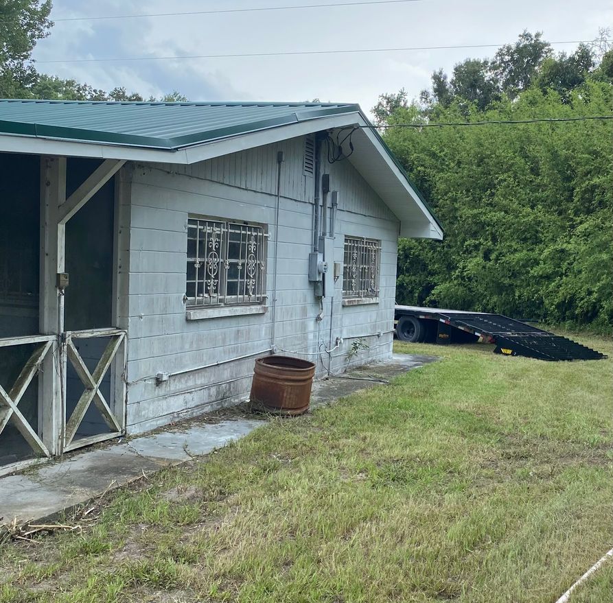 A weathered, white house with a green roof and barred windows, set in a grassy yard with a trailer.