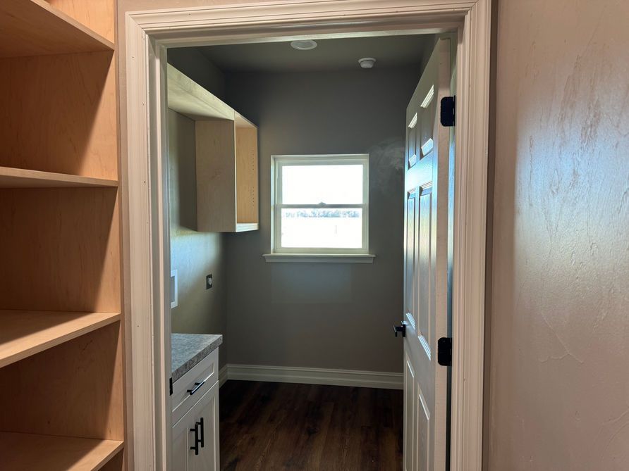 View into a utility room with a window, cabinets, and a door, brown flooring, light tan walls, and an open shelving unit.