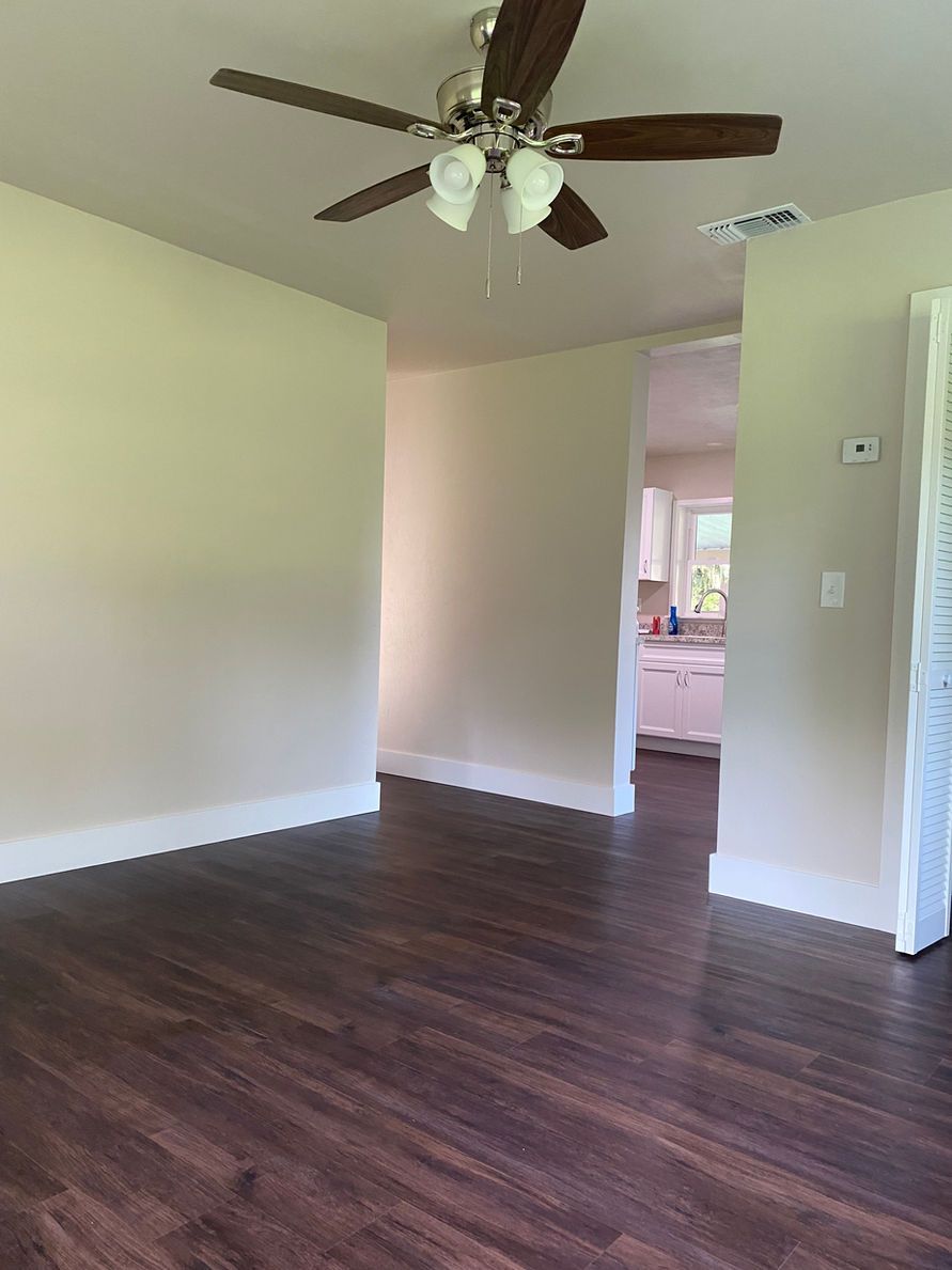 Empty room with brown flooring, off-white walls, and a ceiling fan. A doorway leads to a kitchen.