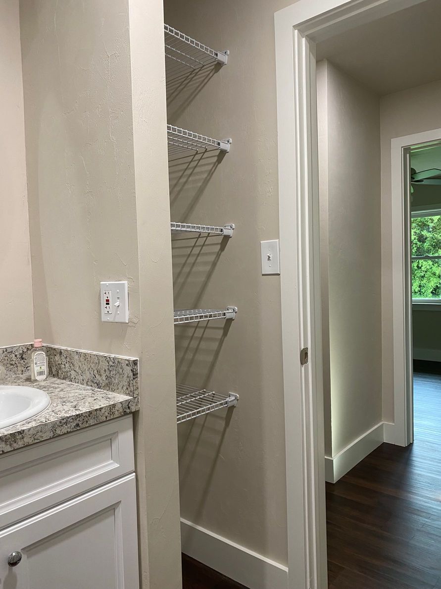 Bathroom with wire shelves next to a doorway leading to a hallway with a window.