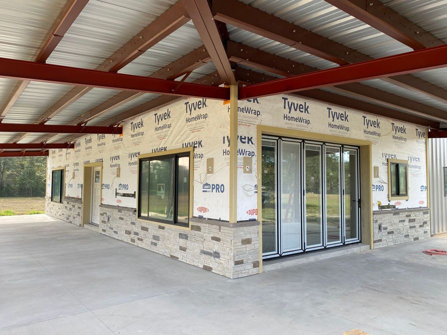 Building under construction; exterior with stone facade, large windows, and Tyvek wrap, under a covered patio.