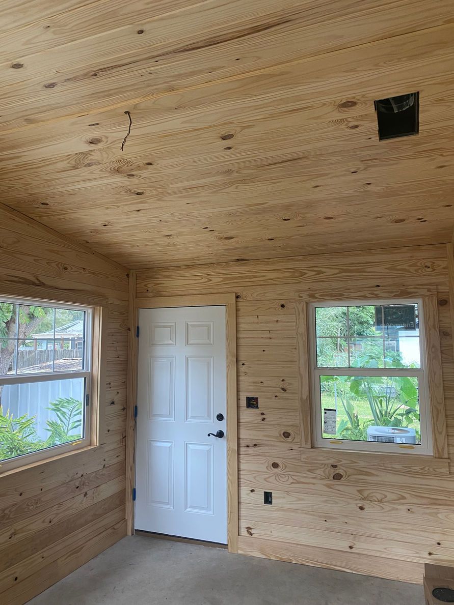 Interior of a small room with wood-paneled walls and ceiling, a white door, and two windows.